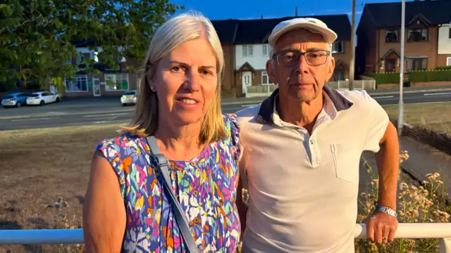 Chris and Pauline Norcup outside in Stoke-on-Trent. Pauline is wearing a floral top with a strap over her shoulder, Chris has a white T-shirt on and a flat cap