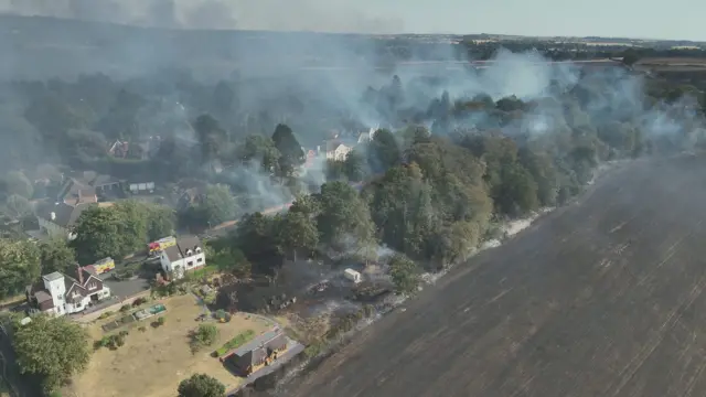 Aerial image showing a scorched field that has turned black while smoke billows from a nearby housing estate.