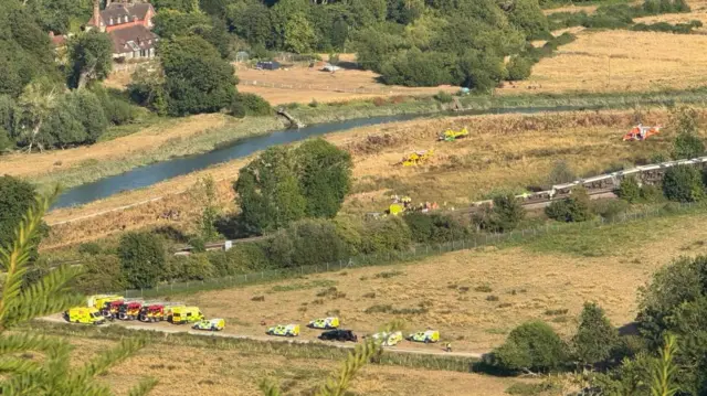 An aerial shot of several emergency services vehicles in a field around a train turned on its side.