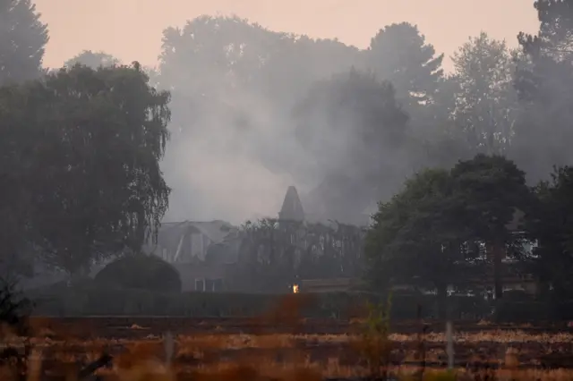 A photo taken from distance showing a heavily damaged house still alight with a glowing flame in the centre of the property. Above the house is a cloud of thick black smoke.