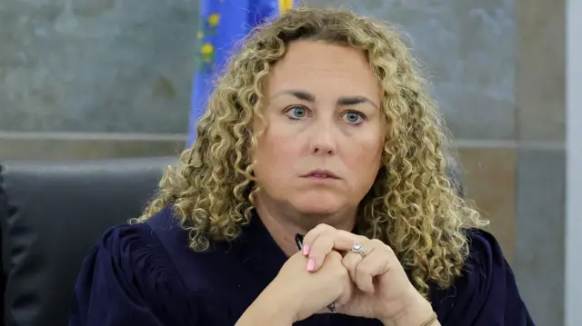 A blonde woman with curly shoulder-length hair clasps her hands together as she looks down from the judge's bench. She has bink nail polish and is wearing a sparkly silver ring on her left ring finger.