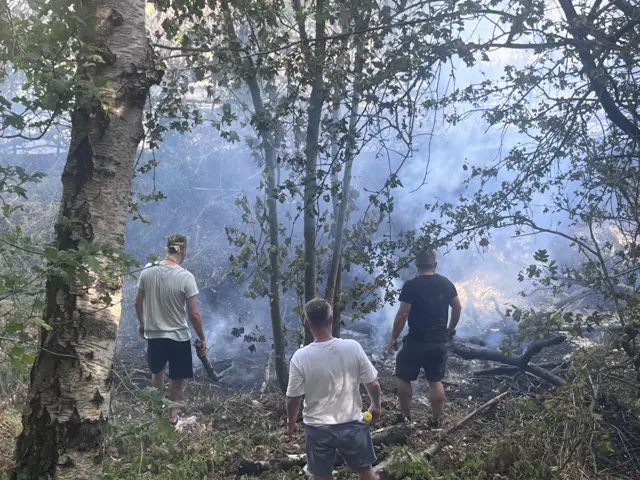A group of men wearing shorts and t-shirts are standing in a wooded areas with smoke billowing in front of fthem.