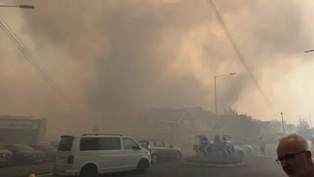 Thick plumes of smoke cover a residential area. Vehicles are queuing around a roundabout as people stand in the street and watch the plume of smoke come down.