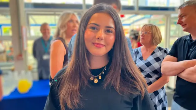Maya has shoulder-length dark hair and is wearing a black top and gold necklace in a bright indoor venue. Several people are gathered in the background near large windows and a refreshments table, with the foreground subject centred and in sharp focus.