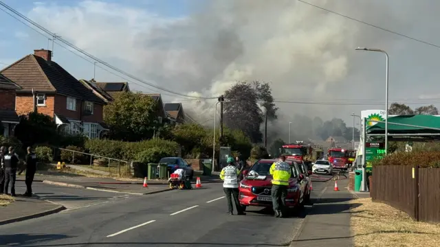 Thick plumes of smoke comes from a row of residential two storey houses. Multiple fire engines and police cars are parked down the street.