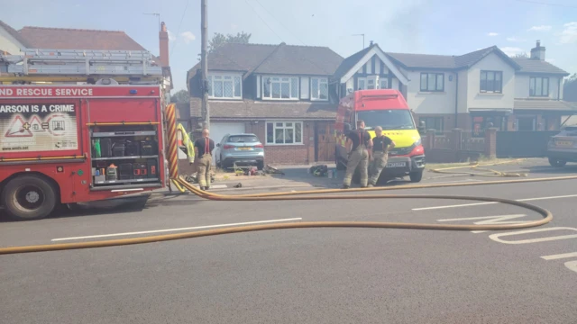 Fire crews are parked along a residential street. One fire fighter is leant against one of the fire vehicles while another fire fighter is stood speaking to him. A brown hose pipe is laid out across the road.