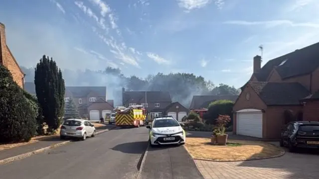 Smoke is pluming above homes with a fire engine and a police car parked in front.