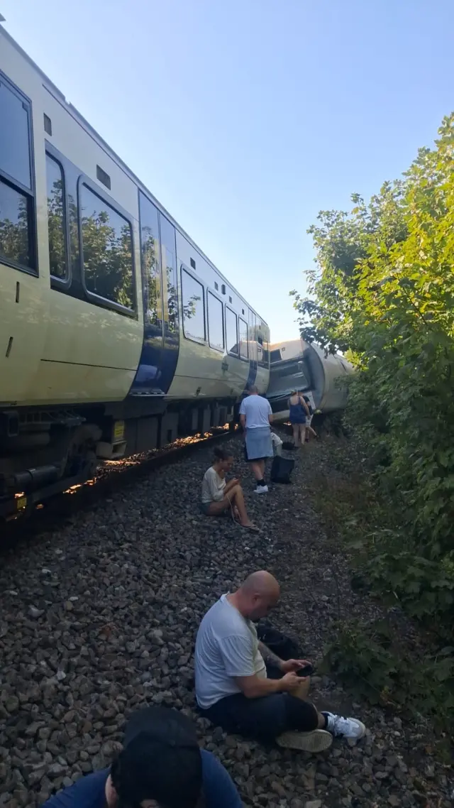 A number of people sat by the side of a railway track. A derailed train is in the background.