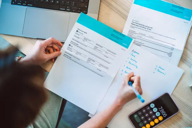 A person writing down notes on a piece of paper while holding an energy bill, the calculator app open on a nearby iPhone