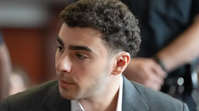 A curly haired man purses his lips and glances to the left, while wearing a grey blazer and a white button up shirt.