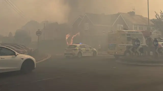 A police car is parked in front of a two storey house which appears to be on fire. Flames are leaping up the side of the property while the air is filled with thick black smoke.
