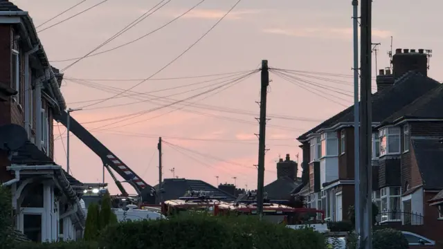 The sun sets as a fire engine attends an affected property. A ladder stretches from the appliance to the upper window of the property.