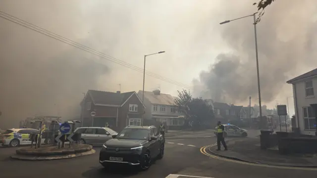 Thick plumes of smoke cover a residential area while police block the entrance to one street.