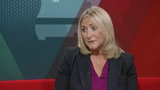 A woman wearing a black suit jacket and a purple shirt is sitting in a tv studio with a red and blue backdrop.