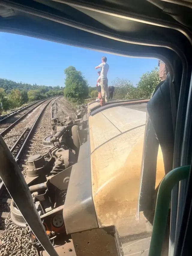 Photo showing a overturned train carriage, with four people standing on top of it, one person's head poking out of an opened door and a buckle in the track