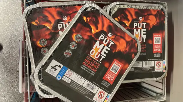 A pile of disposable bbqs on sale. they have foil trays and a picture of red hot coals on the front