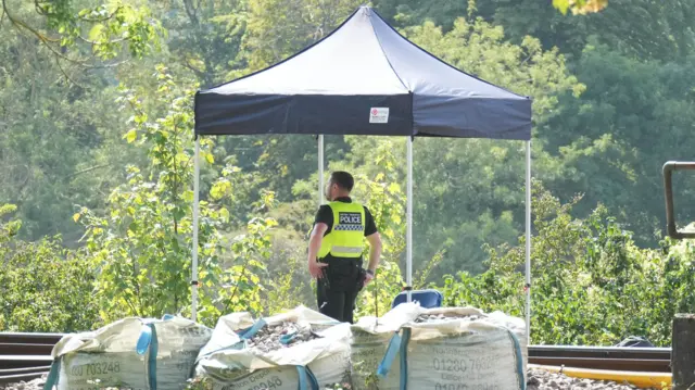 A British Transport Police officer stands beneath a temporary black canopy beside a railway line near Lewes. Bags of track ballast sit in the foreground while trees and vegetation surround the site.