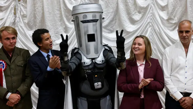 Count binface raises the peace sign with his fingers in a line alongside other candidates