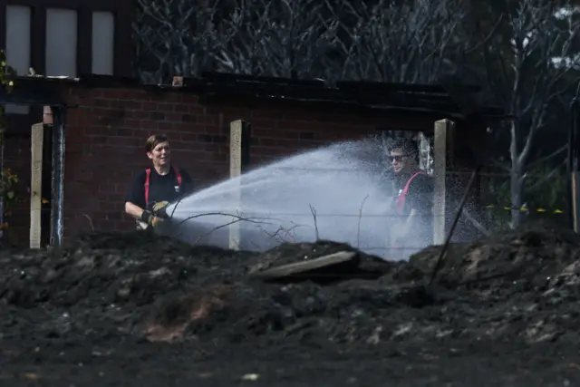 Two firefighters are pictured in the distance past a burnt field. They are stood surrounded by the wreckage of one house while one of them sprays water across the land.