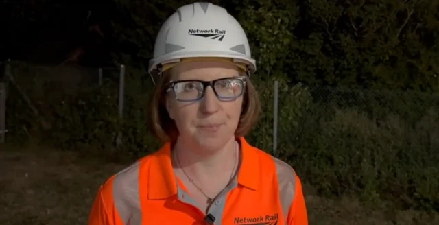 A woman with shoulder-length ginger hair. She is wearing glasses, a white hardhat with "Network Rail" written on it, as well as an orange high vis shirt with "Network Rail" written on it.
