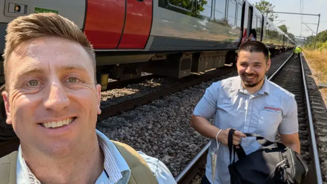 Jon Young has taken a selfie with a member of Greater Anglia staff. Both are walking along the railway line next to the train and wearing pale blue shirts.
