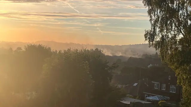 Smoke rises from a fire in the grasslands behind a residential area in the early morning sunlight.