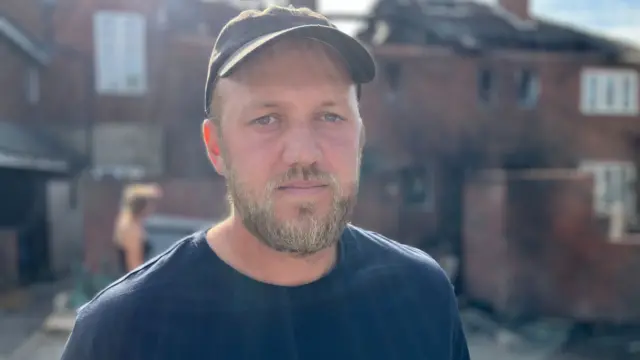 A man wearing a hat and dark t-shirt stands outside a house, which has been damaged by a fire.