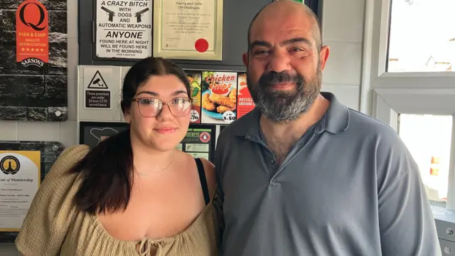 Chris and Maria Christodoulou stood with their arms around each other. Chris is wearing a grey polo top and has a bald head with a dark beard. Maria has long dark hair that is tied back and is wearing a brown top.