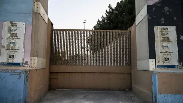 A brown iron gate, closed, leading to the US Embassy compound in Kabul, Afghanistan. To the side, hanging off cement wall, are damaged sign advising people to carry IDs and not take photographs