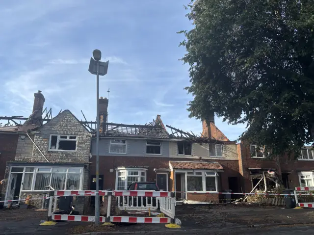 A row of about four or five terraced houses sit partially demolished. Each house is missing its roof with just beams left exposed. Red and white barriers and tape cut the scene off from the public.