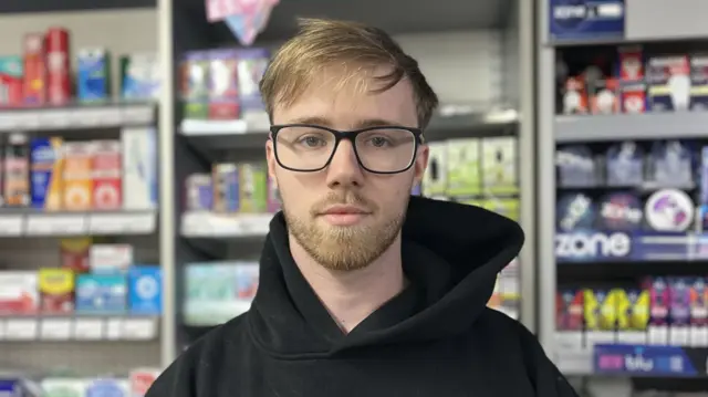 A man with ginger hair wearing a black hoodie and glasses. He is standing behind a counter.
