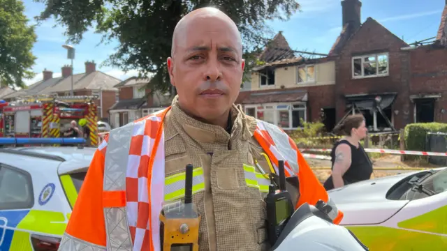 A bald man is wearing a full fire kit which is brown with yellow reflective stripes. He is also wearing an orange and silver hi-vis jacket and is holding a white helmet. Behind him are houses which have barely any roof left because of the fire