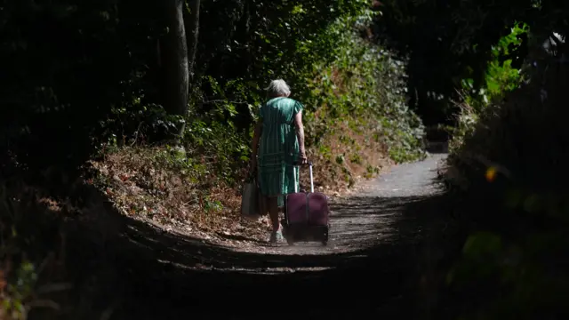 An older woman walking down a country lane away from the camera. She has short white hair and is wearing a green dress. She is holding a bag and dragging a burgundy suitcase