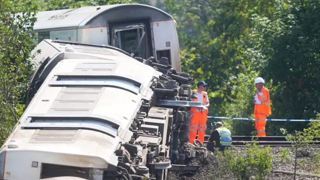 Several passenger train carriages lie on their sides beside a railway line near Lewes. Railway workers in orange high-visibility clothing and a British Transport Police officer are examining the scene next to the overturned vehicles.