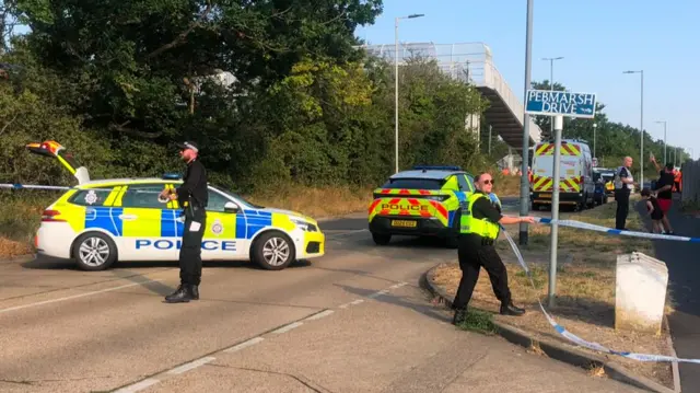 Police officers pull blue and white tape across the road, where various police cars are parked.