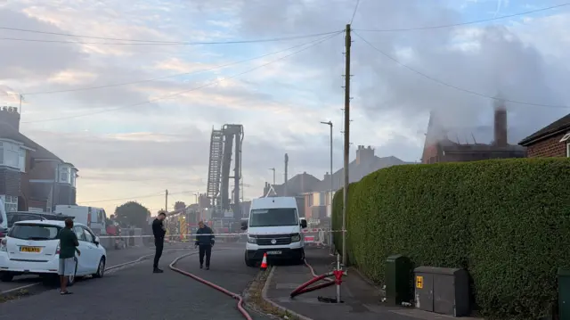 A residential street has been cordoned off as smoke rises from a house destroyed by a fire. People can be seen looking on as firefighters work at the scene. A fire engine with an extended ladder can be seen on the street.