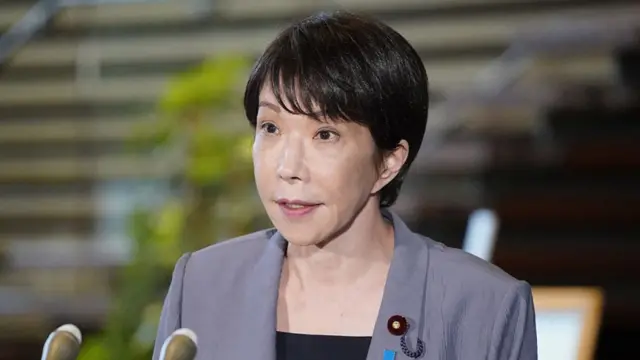 Takaichi speaks in front of two microphones, wearing a gray blazer