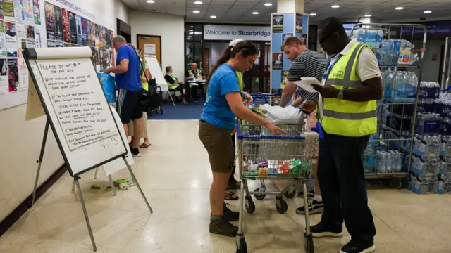 Several people are reaching into a shopping trolley which holds mediation items. Behind them are dozens of bottles of water