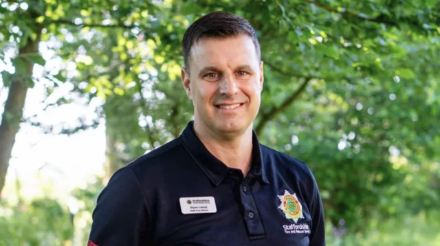 A man wearing fire service uniform is standing outdoors. He is looking directly at the camera and smiling for the photograph. There are trees visible in the background behind him.
