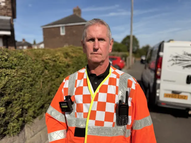 A man in an orange and white checkered firefighter overalls. He has a handheld radio and a video recording device attached to the outfit. He is standing on a pavement with houses and a white van visible behind him.