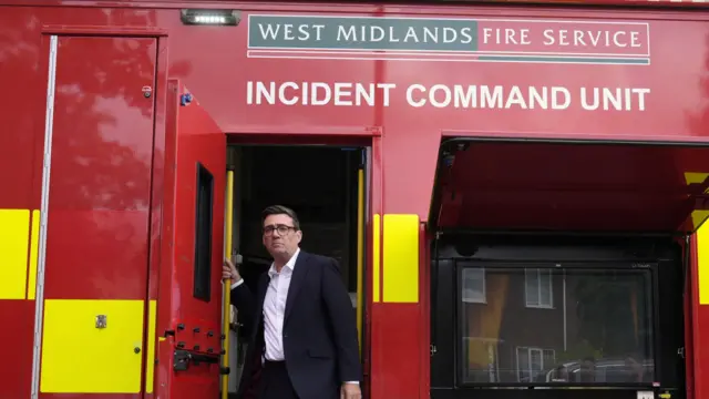 Andy Burnham in a black suit with a white shirt is leaving a mobile unit that is red and yellow. a sign says "West Midlands Fire service incident command unit"
