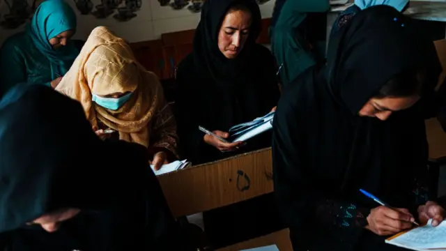 A group of girls wearing veils and abayas sit down at wooden desks taking notes on exercise books