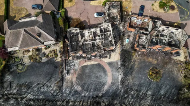 An aerial photo of three houses affected by the fire. The two on the right are completely destroyed and have no roofs. Thier gardens are black and covered in soot. The one on the left still has a roof but their garden is completely black