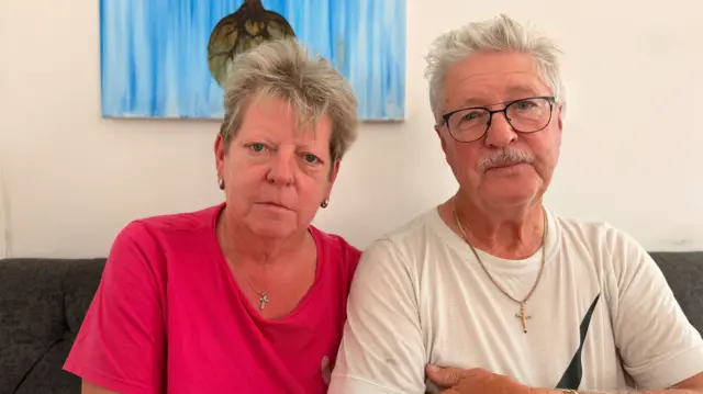 A woman with short grey hair is wearing a pink t-shirt and a man next to her has short grey hair and black glasses and is wearing a white t-shirt