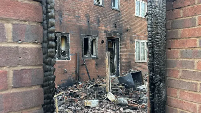 A house which has had its outside blackened by fire. there is burnt piles of rubble outside and pieces of the building. Ground floor windows have been burnt out and have exploded