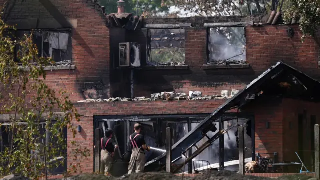 Two firefighters in black t-shirt with red braces and light brown trousers stand in front of a house completely destroyed by fire. It has no roof and the bricks are charred and blackened. Windows are blown out and bits of the building have fallen down