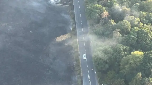 A car travels along a road with a scorched field to one side and a green wooded area to the other.