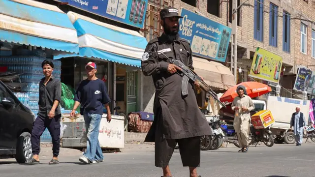 A Taliban official stands in the street holding a gun, as people walk by