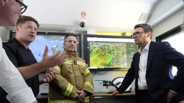 Andy Burnham is standing and talking to several firefighters inside a mobile unit with screens.