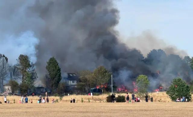 A large blaze, with a huge plume of black smoke engulfs trees and houses. In the foreground are people looking on